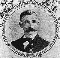 Head and shoulders of a white man with a wide bushy mustache, wearing a bow tie and dark suit with a medal pinned to the left breast. The portrait is surrounded by a circular frame and images of flowers.