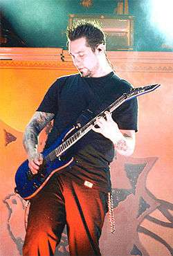 A man with brown hair is wearing a black T-shirt and black trousers while playing on a blue guitar. Tattoos are visible on both of his arms.