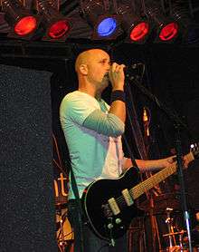 On stage, a man is holding a microphone to his mouth with his right arm while his left hand clasps the neck of a guitar. He is mostly bald headed, staring forward, with five coloured lights above and band equipment obscured, behind.