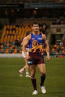 A man in a maroon, blue and gold Australian rules football jersey walks towards the camera on grass