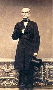 A full-length photograph of a balding man with long sideburns, dressed in a formal frock coat and black tie and holding a black top hat in his left hand at his side