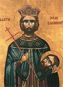 Religious painting of a man in his thirties with a short beard and mustache, wearing a crown and regal robe, holding a cross in his right hand and a severed human head in his left hand.