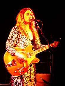 Julia Deans performing on stage, holding a guitar, lit in yellow.
