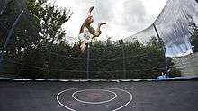 A jumper is mid-jump, upside down, knees tucked, several feet above a large round trampoline surrounded by a high safety net enclosure.