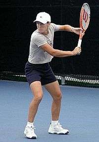 A blond-haired women is wearing a neon-pink shirt and white skirt, and is reaching to hit a one handed backhand
