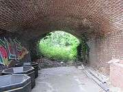 A barrel-vaulted brown brick tunnel approximately 10 metres long & 5 metres wide leads to a lush green clearing. The tunnel has graffiti & is being used to store large black bins.