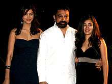 Bearded man in white, flanked by two young women in black dresses