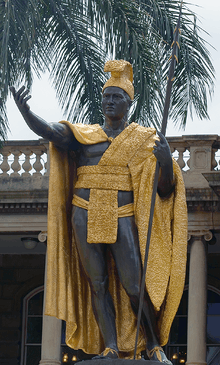 An impressive statue of a dark-skinned man, richly dressed in native garb.  One arm is extended, as if in greeting, and he holds a spear, butt end grounded, in the other.