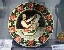 A painted plate, surrounded by cabbage roses, has a bearded man painting an umbrella stand with cabbage roses. At the bottom a caption reads "Karel Nekola 1857-1915".