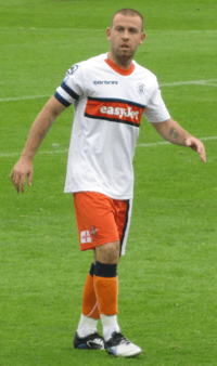 A young man with close-cut dark hair walks to the viewer's right, attired in a white and orange soccer kit. His eyes look to the photographer's left and a blue and white captain's armband is prominent on his right arm.
