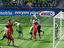 Several players in red uniforms and green uniforms in front of goal with a goalkeeper in black leaping with his hand hitting a soccer ball
