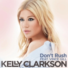 An image of a blonde looking aside against a pinkish background. A blue shirt is draped around her shoulder. Below her, the words "Don't Rush feat. Vince Gill" is written in grey color. Below these words, the word "Kelly Clarkson" is written in black capital letters.