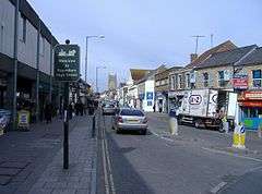 Street scene showing shops on left and right, with cars and vans on road. On the left hand pavement is a sign saying welcome to Keynsham high street.