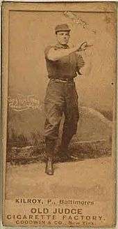 A baseball card of a man in a dark baseball uniform wearing a matching baseball cap and holding his hands in the air in front of his chest.
