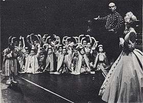 Black and white photo of a theatrical scene: a middle-aged man with a shaved head and imposing presence has his right arm extended to introduce a group of children in Asian dress to a woman in a crinoline dress and bonnet in the foreground at right, who is partially turned upstage.  The children are mostly kneeling and have their arms raised in greeting; one child (probably Crown Prince Chulalongkorn) stands and bows.