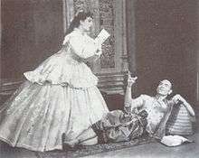 A man with a shaven head, wearing Asian dress, reclines on the floor and gestures at a woman in 19th century dress, who is writing, apparently at the man's dictation.