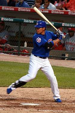 A man in a blue batting helmet with a "C" on it, blue baseball jersey, white pinstriped pants, and a shin-guard on his right shin stands at home plate holding a baseball bat in a left-handed batting stance with his right foot lifted off of the ground.