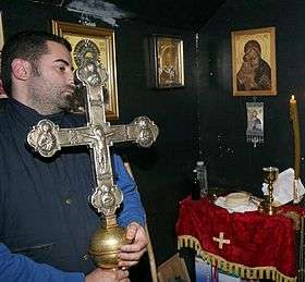 Photograph of a man in his thirties holding a large silvery cross in a room on whose walls religious paintings are hung.