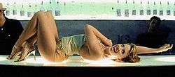 A woman with dark blonde hair is laying on a glass bar counter, which features a blue colour scheme. The woman is dressed in a pale green-tinged jersey top, gold lamé hotpants, and golden high-heels