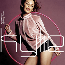 A woman dressed in a rose-coloured jacket and black hot-pants is striking a pose in front of a dark purple background with a pink hoop in her hand. The word "Kylie" is written in front of her in a large white font.