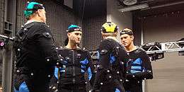 Four actors talking in motion capture suits (black full-body suits, and a small cap upon the head, with small white balls), in a studio.