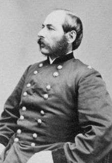 Head and torso of a white man with bushy sideburns connecting to his mustache, wearing a plain double-breasted military jacket.
