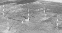 Aerial view of five tall Adcock antenna towers standing on flat terrain; four are arranged in a square, and the fifth one is at the center