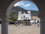 View through an arch onto a paved square with a fountain in the center and a white church on the far side