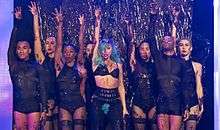 A group of people standing with their right hand raise up. They are decked in black garments with bare legs. Central to them is a woman who has blue hair and tufts of hair from her underarms and at her pubic area.