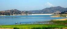 A light blue, broad lake is formed by a dam seen in the distance in a wide valley between arid hills
