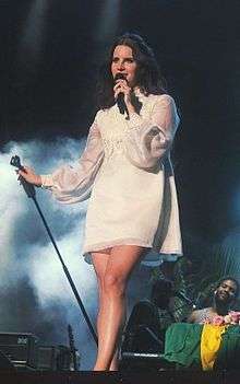 A light-skinned, brown-haired young woman wearing a white long-sleeved dress is standing on a stage singing into a microphone