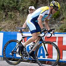 A cyclist in a blue and white jersey with yellow trim, bereft of any writing on it. He is riding out of the saddle as he climbs a hill, as a spectator watches on from behind a barricade.