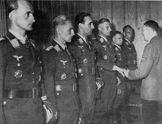 Six men all wearing military uniforms and decorations standing in row. The third man from the far right is shaking hands with another man.