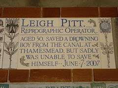 Almost identical to the plaque to Amelia Kennedy, a tablet formed of five tiles of varying sizes, bordered by yellow and blue flowers in an art nouveau style and decorated by two stylised salmon. The tablet reads "Leigh Pitt, reprographic operator, aged 30, saved a drowning boy from the canal at Thamesmead, but sadly was unable to save himself June 7, 2007".
