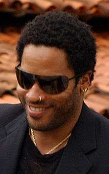 A dark skinned man with sunglasses smiles. His hair is short and tightly curled. He wears a dark suit jacket and sports stubble. The man has hooped rings in his left ear and left nostril.