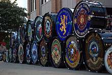 Bass drums, each marked with a distinctive logo of a fire department, are stacked in front of a building