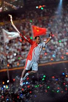 An Asian man in red-and-white athletics shirt and shorts, and wearing sneakers, is suspended by wires in the air while holding a lit torch in his right hand. In the background, a large crowd in a stadium can be seen, as well as two blurred flags hoisted in flagpoles.