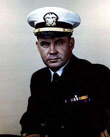 A man in a dark suit and tie, with a white peaked cap. Hears wears the two gold stripes of a lieutenant on his sleeve, and ribbons including those of the Distinguished Service Cross and Silver Star.