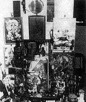 Various portraits and statuettes are arrayed on a small table and the wall it is against.  A small round mirror hangs above this collection of religious items.