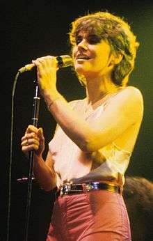 Ronstadt, in the summer of 1978, sings in concert dressed in a camisole top and pants with medium-length hair, clutching the microphone on a stand with both hands.