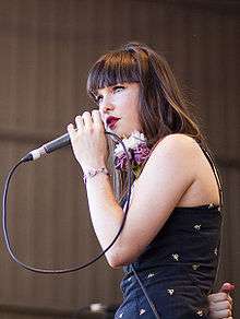 A 19-year-old woman is shown in upper body, left profile. She holds a microphone close to her lips using her left hand, the cord is looped back to her hand. Her right arm is clenched behind her back. She wears dark brown hair, her dark blue dress has a pattern of small yellow-white logos, and a large flower is near her shoulder. Her left wrist has a bracelet and her eyes are directed to her left. Beyond her the wall of the venue is blurred.