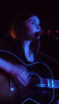 18-year-old Mitchell is shown in an upper body shot. She plays a guitar while singing into a microphone. She is tinged blue-white due to lighting and her right hand, which is strumming the guitar strings, is blurred.