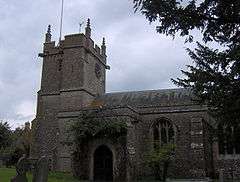 Stone building with square tower at left hand end. In the foreground are trees and gravestones.