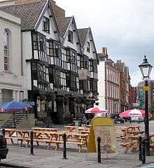 A seventeenth-century timber-framed building with three gables and a traditional inn sign showing a picture of a sailing barge. Some drinkers sit at benches outside on a cobbled street. Other old buildings are further down the street, and in the background part of a modern office building can be seen.