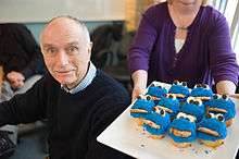 A Caucasian man in his early 80s, wearing a dark blue sweater, and looking directly into the camera.  On his right is a plate of cupcakes decorated like Cookie Monster.