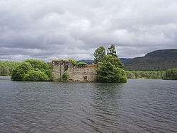 A small wooded islet stands in a lake. A large ruined stone wall with a door and window sits amongst the trees. Green conifer-clad hills lie beyond the islet under leaden grey skies.