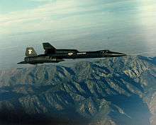 Sideview of black jet aircraft overflying mountain towards right of photo.