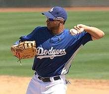 A man in a blue baseball jersey with "DODGERS" on the chest with a baseball glove on his right hand prepares to throw a baseball with his left hand.