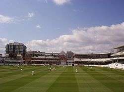 A view of a cricket ground with blue sky and some clouds, the stands surrounding the pitch are mainly unoccupied, to the left in the background is a large building