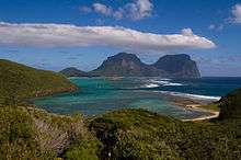 View overlooking a sheltered shallow bay, with a reef extending out to two bald peaks in the background
